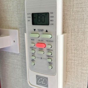 May include: A white air conditioner remote control with a digital display showing 80 degrees Fahrenheit. The remote has buttons for fan speed, temperature, and other settings. The brand logo "CL" is visible.