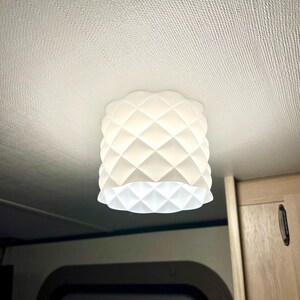 May include: A white, geometric-patterned ceiling light fixture. The light has a cylindrical shape with a diamond pattern and a scalloped edge at the bottom. The light is mounted on a white ceiling.