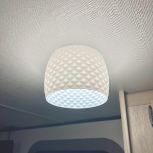 May include: White, geometric-patterned pendant light fixture. The light has a rounded, bell-like shape with a textured surface. The light is hanging from a white ceiling. The bottom of the light is illuminated, casting a soft glow.