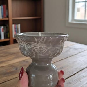 May include: A grey ceramic pedestal bowl with a flared rim and a rounded base. The bowl has a white, etched floral design around the rim. The bowl is held in a person's hand, with a wooden table and a bookshelf in the background.
