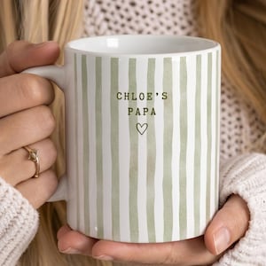 May include: White ceramic mug with vertical green stripes. The mug has the text "Chloe's Papa" and a heart symbol. The mug is held by a person wearing a white sweater.