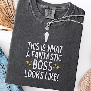 May include: Dark gray Comfort Colors t-shirt with the text "THIS IS WHAT A FANTASTIC BOSS LOOKS LIKE!" in white. A silver necklace with a pendant is draped across the shirt. The shirt has a crew neck and short sleeves.