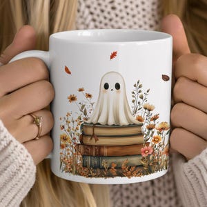 May include: A white ceramic mug featuring a graphic illustration of a small, pale ghost sitting atop a stack of four vintage-style books. The scene is surrounded by autumn-themed wildflowers in shades of orange, yellow, and brown with falling leaves.