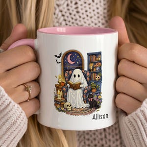 May include: A white ceramic mug with a pink interior and handle, featuring a printed illustration of a ghost reading a book in an armchair, surrounded by a bookshelf, a black cat, skulls, and candles, with the name Allison printed below the image.