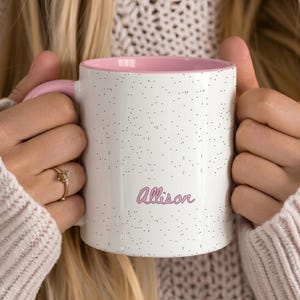 May include: A white ceramic mug with a speckled black pattern, a pink interior, and a pink handle. The name Allison is printed on the front in a pink cursive script font. The mug features a smooth finish and a cylindrical shape for holding beverages.