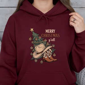 May include: Burgundy hoodie with a Christmas graphic. The design includes a Christmas tree, cowboy hat, and boots, with the text "MERRY CHRISTMAS y'all" in gold. The hoodie has a drawstring hood and a front pocket.