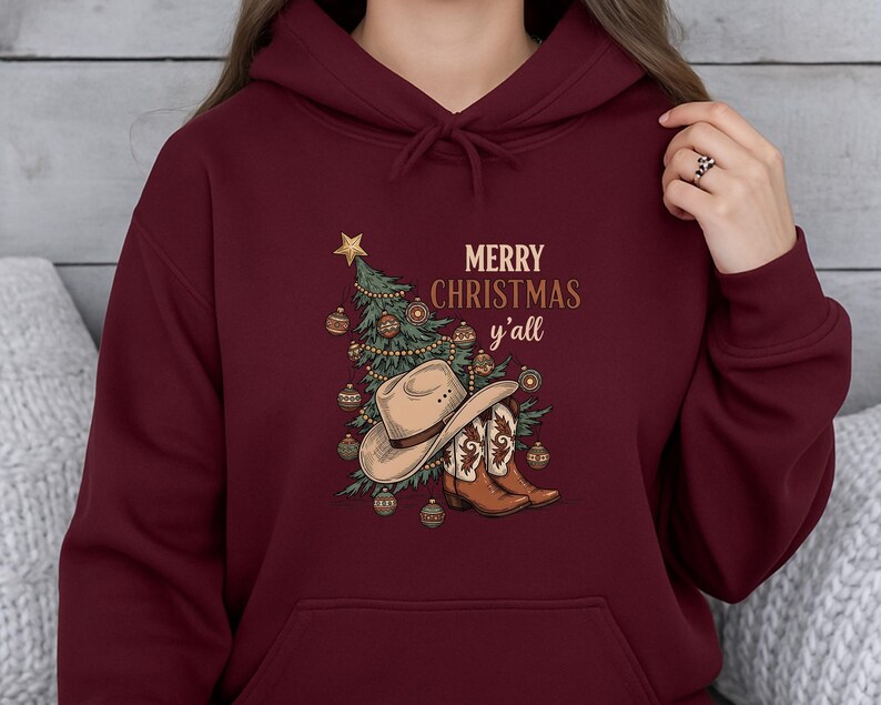 May include: Burgundy hoodie with a Christmas graphic. The design includes a Christmas tree, cowboy hat, and boots, with the text "MERRY CHRISTMAS y'all" in gold. The hoodie has a drawstring hood and a front pocket.