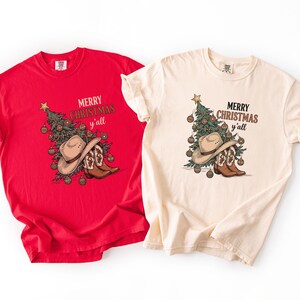 May include: Two t-shirts, one red and one cream, with a festive Christmas design. The design features a Christmas tree, cowboy hat, and boots, and the text "Merry Christmas y'all".