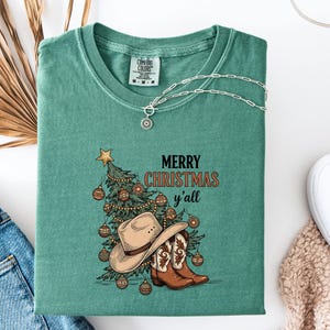 May include: A teal Comfort Colors t-shirt with a Christmas graphic. The graphic features a decorated Christmas tree, cowboy hat, and boots, with the text "MERRY CHRISTMAS y'all". A silver necklace with a pendant is draped on the shirt.
