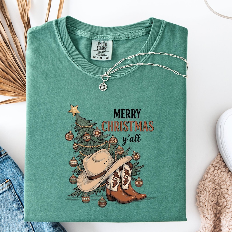 May include: A teal Comfort Colors t-shirt with a Christmas graphic. The graphic features a decorated Christmas tree, cowboy hat, and boots, with the text "MERRY CHRISTMAS y'all". A silver necklace with a pendant is draped on the shirt.