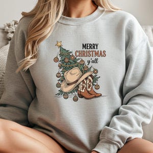 May include: A light grey sweatshirt with a Christmas tree, cowboy hat, and boots design. The words "MERRY CHRISTMAS y'all" are printed above the graphic in brown and black. A casual, festive garment for the holiday season.