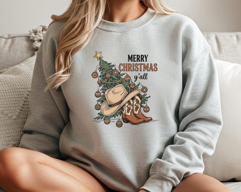 May include: A light grey sweatshirt with a Christmas tree, cowboy hat, and boots design. The words "MERRY CHRISTMAS y'all" are printed above the graphic in brown and black. A casual, festive garment for the holiday season.