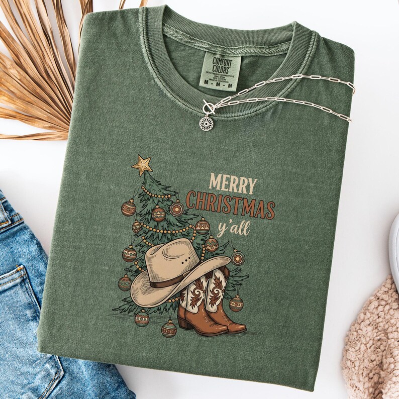 May include: An olive green t-shirt featuring a Christmas tree, cowboy hat, and boots graphic. The text "MERRY CHRISTMAS y'all" is printed on the front. A silver necklace is draped across the shirt.