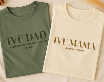 May include: Two t-shirts, one olive green and one cream-colored, are displayed. The olive green shirt reads "IVF DAD" with the text "Married to the toughest warrior I know." The cream shirt reads "IVF MAMA" with the text "Toughest warrior."