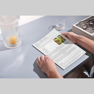 May include: A person holding a printed document with text and an image of colorful flowers. The document is on a gray surface, with a glass of water and a decorative glass container with a yellow object inside.