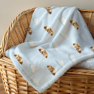 May include: A light blue baby blanket with a repeating pattern of sleeping puppies and white clouds. The blanket is made of a soft, plush material and is draped over a wicker basket.