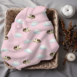 May include: A pink blanket featuring a pattern of sleeping puppies and white clouds. The blanket is folded inside a wicker basket. The scene is set against a neutral backdrop.