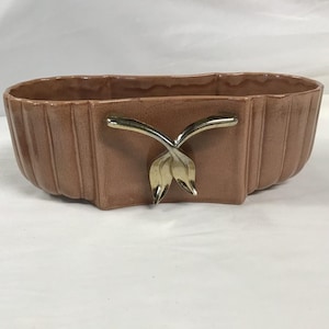 May include: A vintage, oblong planter in a muted brown hue. The ceramic planter features vertical ribbed detailing and a gold-toned, stylized leaf design on the front. The planter is a decorative item for displaying plants or flowers.