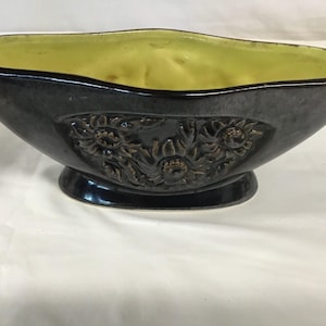 Könnte beinhalten: Dunkelgrauer Keramik-Blumentopf mit erhabenem Blumendesign. Der Innenraum ist ein leuchtendes Gelbgrün. Der Pflanztopf hat einen Sockel und eine sanft geschwungene Form, geeignet für die Präsentation von Pflanzen oder als dekoratives Stück.