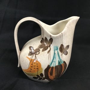 May include: Cream-colored ceramic pitcher with a curved handle and spout. The pitcher is decorated with brown leaves, an orange gourd, and a blue bottle design. The pitcher has a speckled finish.