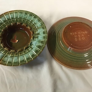 Puede incluir: Juego de ceniceros de cerámica de dos piezas. La pieza superior es de color verde oscuro con un patrón de cuadrícula y un cuenco central. La pieza inferior es marrón y verde con el texto "RED WING USA 884".