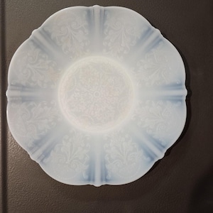 May include: A white, scalloped-edge plate with a delicate floral pattern. The plate has a soft, opalescent glow and a detailed design in the center. The plate is likely made of glass or ceramic and is suitable for serving food.