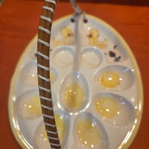 May include: Oval-shaped, white ceramic deviled egg serving dish with yellow trim and a woven handle. The dish has egg-shaped indentations, some filled with yellow egg yolks. Two yellow duckling illustrations are painted on the dish.