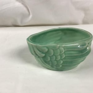 Könnte beinhalten: Eine kleine, oval geformte, hellgrüne Keramikschale. Die Schale hat ein strukturiertes Design an den Seiten, das Federn oder Blättern ähnelt. Der Innenraum ist glatt und die Schale steht vor einem weißen Hintergrund.