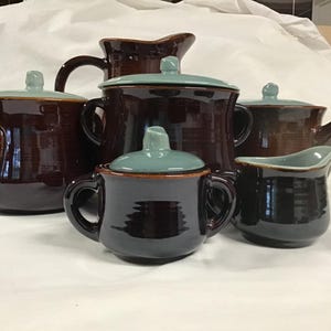 Könnte beinhalten: Eine Sammlung brauner Keramik-Servierteile mit tealfarbenen Deckeln und Akzenten. Das Set umfasst eine Teekanne, eine Zuckerdose, einen Sahnekännchen und Krüge. Die Teile haben eine glänzende Oberfläche und sind auf einer weißen Oberfläche angeordnet.