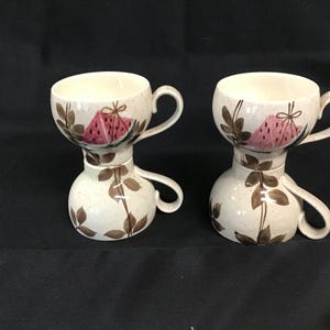 Könnte beinhalten: Zwei Keramik-Teetassen. Die Tassen sind cremefarben mit einer rosa Wassermelonen-Scheibe und braunen Blattakzenten. Jede Tasse hat einen Henkel und einen bauchigen Sockel mit einem Henkel.