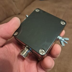 Puede incluir: Un dispositivo electrónico negro y cuadrado con un conector plateado y una tuerca de mariposa. El dispositivo tiene cuatro tornillos en las esquinas y se sostiene en una mano.