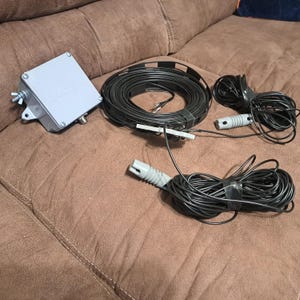 May include: A grey metal box with the text "DATEX" and several black coiled cables with grey connectors. The cables are bundled together with black tape. The items are on a brown surface.