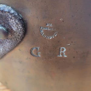 May include: Close-up of a bronze-coloured metal object with the embossed letters "G" and "R". Above the letters is a circular emblem with the text "DIL PADEMA HANNOVER". The object has a slightly textured surface.