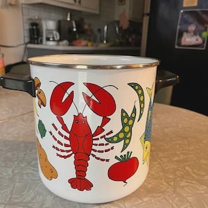 May include: White enamel cooking pot with black handles and a silver rim. The pot is decorated with colorful illustrations of a red lobster, vegetables, and fish. A charming kitchenware item.