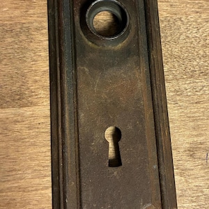May include: A rectangular, dark brown, weathered metal door plate featuring a central circular opening for a knob and a lower keyhole. The plate has a raised border and four small screw holes, one at each corner, showing signs of oxidation.