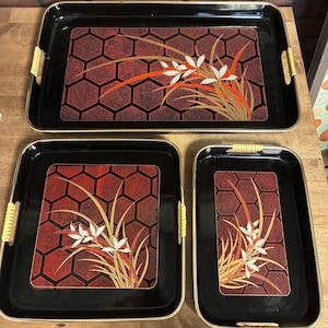 May include: A set of three black serving trays with gold trim and woven handles. Each tray features a red and black geometric pattern with a floral design in white, orange, and gold.