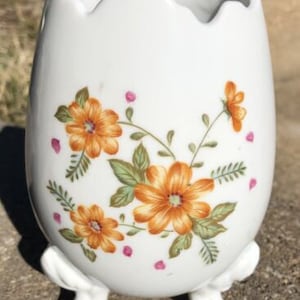 May include: White ceramic egg-shaped vase with three legs and a floral design featuring orange and pink flowers.