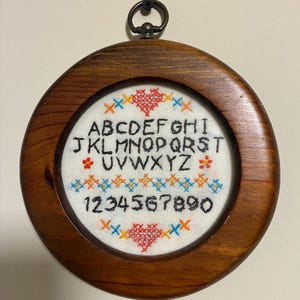 May include: A circular wooden frame containing white fabric with black embroidered letters of the alphabet and numbers zero through nine. The embroidery features red heart motifs and small blue and orange cross-stitch patterns above and below.