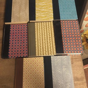 May include: A collection of vintage books with decorative covers. The covers feature various patterns, including floral, geometric, and marbled designs in colors like blue, yellow, red, and cream. The spines are black or brown.