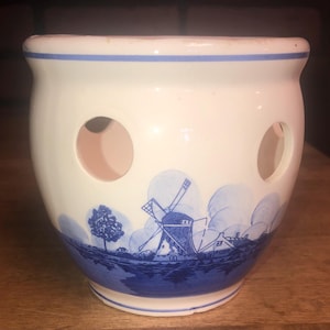 May include: A white ceramic onion keeper with three circular holes and a blue rim. The keeper features a blue and white landscape design with a windmill, trees, and houses. The interior is a light pink color.