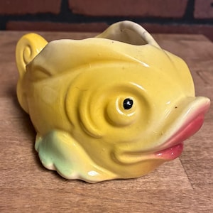 May include: A yellow ceramic creamer shaped like a fish with a glossy finish. The fish features a prominent black eye, pink-tinted lips, and light green accents on the lower fins. The top of the fish is open, forming a spout for pouring liquids.