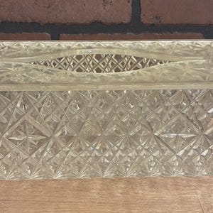 May include: A clear, rectangular tissue box with a diamond-cut pattern. The box has a rectangular opening on the top for easy access to tissues. The box is made of a clear material, allowing the tissues inside to be visible.