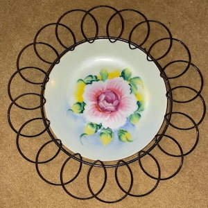 May include: A decorative plate with a hand-painted pink rose design, surrounded by a dark brown metal wire frame. The plate has a cream-colored background with yellow and green accents. A vintage-style piece for display.