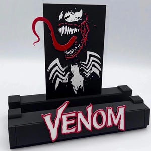 May include: Black phone stand featuring the word "Venom" in white with a red outline. The stand holds a black card with a white and red graphic of Venom's face. The design includes a long red tongue and sharp teeth.