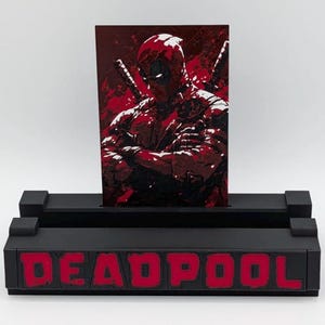May include: A black display stand with the word "DEADPOOL" in red lettering. The stand holds a red and black graphic of Deadpool with crossed swords. The artwork is a comic book style illustration.
