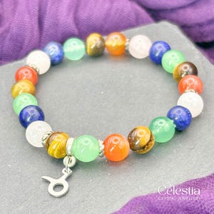May include: A colorful beaded bracelet with a silver charm. The bracelet features a variety of gemstone beads in shades of white, blue, green, orange, and brown. The charm is a Taurus symbol. The bracelet is on a gray surface with a purple background.