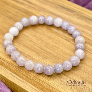 May include: A light purple beaded bracelet with round beads. The bracelet is made of polished stones and is displayed on a wooden surface. The brand name "Celestia Crystal Jewellery" is visible in the bottom right corner.