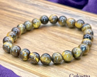 Puede incluir: Un brazalete de piedras preciosas de ojo de tigre pulidas. Las cuentas redondas, de color marrón dorado, tienen rayas marrones oscuras y negras. El brazalete se exhibe sobre una superficie de madera. Las cuentas miden aproximadamente 1 cm de diámetro.