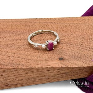 May include: A silver-toned ring featuring a central oval-shaped deep pink gemstone flanked by two small white pearls and two tiny clear stones on either side. The band is adjustable with an open gap at the back, showcasing a polished metallic finish.