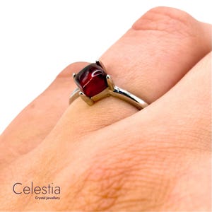 Peut inclure: Une bague en argent ornée d'une pierre précieuse carrée, rouge foncé. La bague est sertie de quatre griffes et le bracelet est en argent poli. Le mot "Celestia" est visible en bas à gauche, avec "Crystal Jewellery" en dessous.